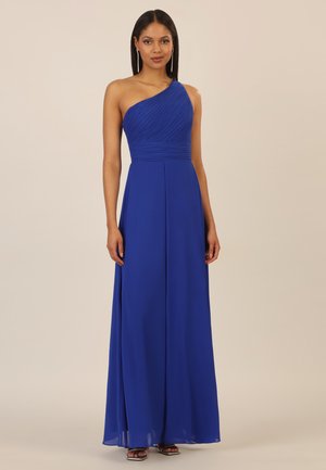 Frau in einem bodenlangen, einseitig schulterfreien Abendkleid in königsblau, die vor einem schlichten beigen Hintergrund steht.