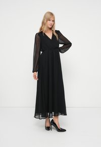 Frau steht mit der Hand in der Hüfte, trägt ein langes schwarzes Kleid mit transparenten Ärmeln und schwarze High-Heels vor einem weißen Hintergrund.