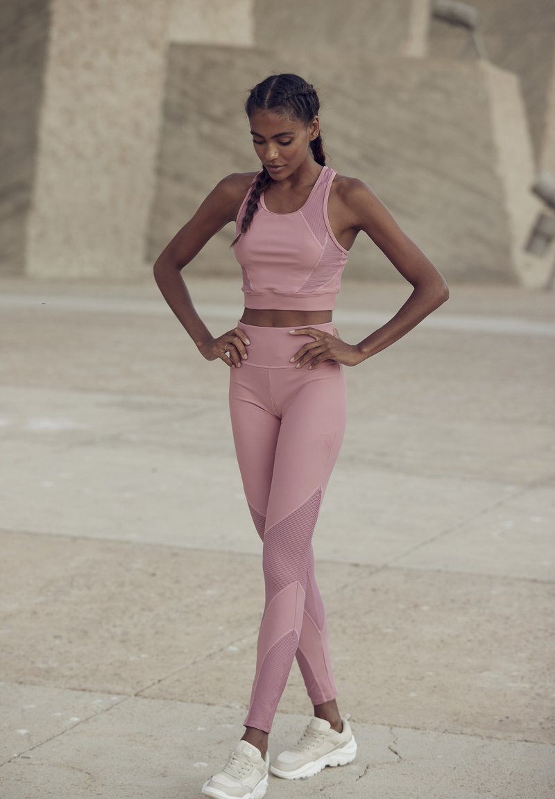 Light pink athletic crop top and leggings with textured accents, paired with white chunky sneakers, set against a neutral background.
