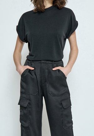 Mujer con una camiseta negra de manga corta metida en unos pantalones cargo negros de talle alto, con las manos en los bolsillos.