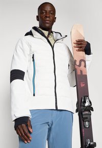 Biela zateplená lyžiarska bunda s čiernymi akcentmi a modrými zipsami, v kombinácii so svetlomodrými lyžiarskymi nohavicami a snowboardom s grafickými dizajnmi.