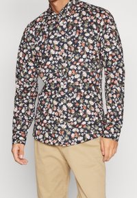 Chemise à manches longues florale en bleu marine foncé avec des fleurs blanches, roses et orange. Col boutonné et tissu à texture lisse.