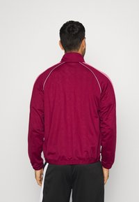 Körsbärsröd zip-up jacka med hög krage och vita pipingdetaljer. Tillverkad av ett mjukt tyg med ett subtilt mönster. Elastisk nederkant och ärmslut.