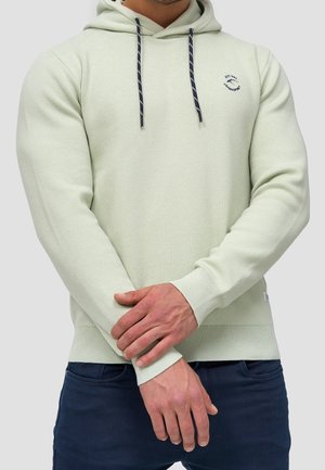 Mann trägt hellgrünen Kapuzenpullover mit dunkelblauen Kordeln, verschränkt die Hände vorne, kombiniert mit dunkelblauer Hose.