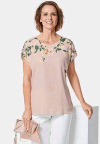 Helles rosa Blusenshirt mit floralem Druck am Ausschnitt, kurzen Ärmeln und einem fließenden Stoff. Kombiniert mit weißen Hosen und einer metallischen Clutch.