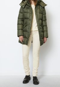 Olijfgroene donzen jas met capuchon, ritssluiting en drukknopen, gecombineerd met lichtbeige broek en zwarte chunky schoenen.