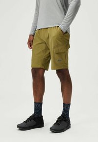 Cargo-Shorts in Olivgrün, aus strapazierfähigem Stoff. Verfügen über mehrere Taschen und ein Patch-Label. Getragen mit schwarzen Sneakers und gemusterten Socken.