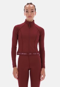 Vīnsarkanā sporta jaka ar rāvējslēdzēja aizdari, garām piedurknēm un piegulošu siluetu. Izceļas ar kontrastējošu baltu "BAKER SPORT" joslu viduklī.