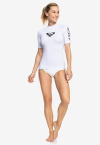 Vit rashguard med korta ärmar och en svart logotyp på bröstet och armen. Tillverkad av stretchigt, slitstarkt material som ger en figurnära passform.