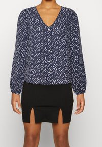 Blusa navy a maniche lunghe con motivi astratti bianchi, scollo a V e chiusura con bottoni. Abbinata a una mini gonna nera con spacchi laterali.