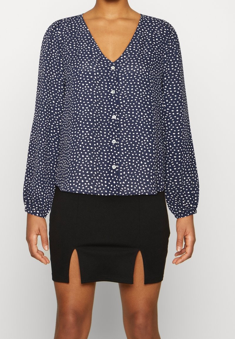 Blusa navy a maniche lunghe con motivi astratti bianchi, scollo a V e chiusura con bottoni. Abbinata a una mini gonna nera con spacchi laterali.