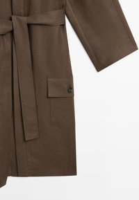 Manteau marron à manches longues avec une ceinture en tissu nouée à la taille et une poche à rabat rectangulaire avec un bouton noir sur le côté.