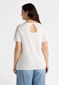 Camiseta blanca de manga corta con un corte en forma de llave en la espalda, hecha de una tela suave. Combinada con pantalones de mezclilla azul claro.