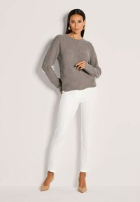 Frau steht vor einer hellen Wand, trägt einen strukturierten grauen Pullover, weiße eng anliegende Hosen, nudefarbene High Heels und geometrische Ohrringe aus Gold.