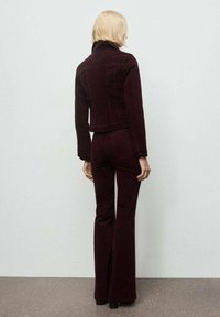 Giacca in corduroy bordeaux e pantaloni a zampa, con un design aderente e vita alta, mostrati da dietro su uno sfondo tenue.