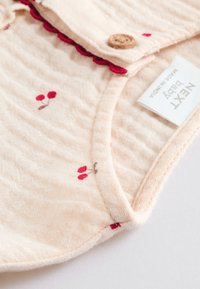 Tissu pêche pâle avec imprimés de cerises rouges, bord festonné et détail de bouton en bois. L'étiquette indique "Next Baby." Surface douce et texturée.