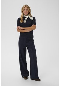 Moteris, turinti šviesius plaukus, apsirengusi tamsiai mėlynu trumpomis rankovėmis megztiniu viršumi, plačiomis tamsiai mėlynomis kelnėmis, ruda avalyne ir kremine skarele su mėlynomis detalėmis, surišta ant kaklo.