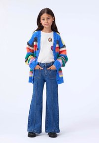 Multicolored fuzzy cardigan featuring red, blue, and green stripes, worn over a white shirt with embroidered patches, paired with flared jeans.