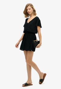 Vestido corto negro con mangas acampanadas y escote en V, combinado con sandalias planas negras y un bolso de mano negro trenzado. Textura de tela suave.