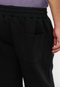 Άτομο που φοράει μαύρο παντελόνι με ελαστική μέση και πίσω τσέπη, εμφανιζόμενο από τη μέση έως το μέσο του μηρού, με το χέρι χαλαρό στο πλάι.