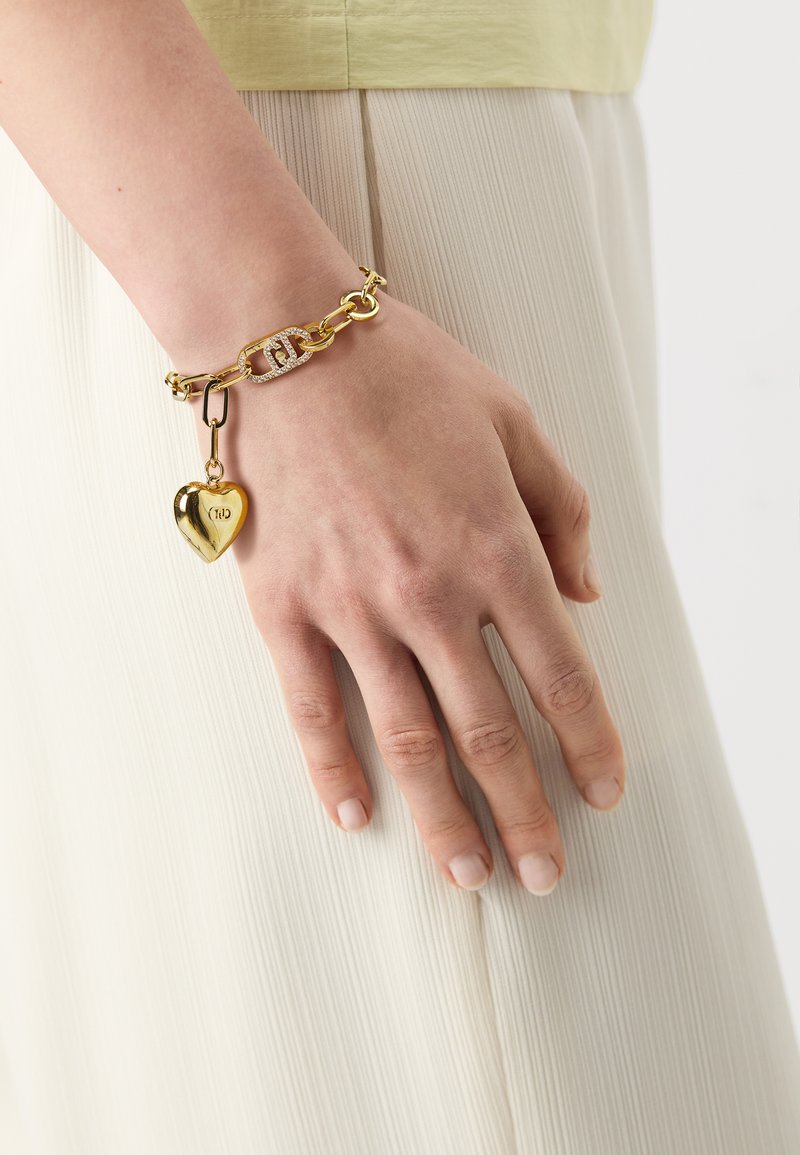 Gullkjedearmbånd med hjerteanheng og en link med krystalldetaljer. Armbåndet har en glatt metallisk tekstur og en dristig form.