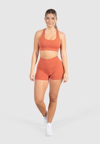 Giovane donna in piedi, che indossa un coordinato di reggiseno sportivo e pantaloni corti arancioni, scarpe da ginnastica bianche, su uno sfondo grigio chiaro uniforme.