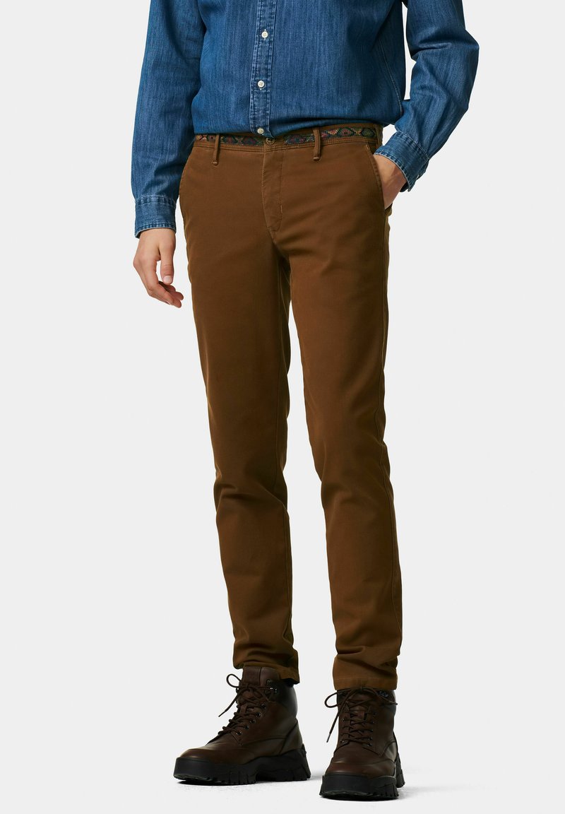 Braune Slim-Fit-Hosen mit einer glatten Textur, kombiniert mit eleganten, dunkelbraunen Leder-Schnürstiefeln. Ein blaues Jeanshemd mit Knöpfen rundet das Outfit ab.