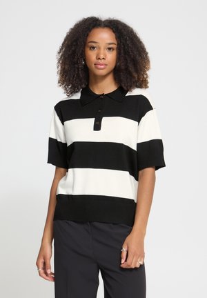 Mujer con cabello rizado, viste una camisa polo de manga corta a rayas negras y blancas y pantalones negros sobre un fondo liso.
