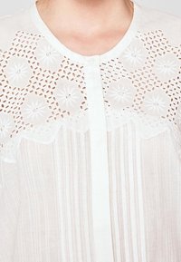 Witte blouse met een knoopsluiting aan de voorkant, voorzien van een bloemenborduursel op de schouders en delicate kanten afwerking. Verticale gestreepte textuur over de hele blouse.