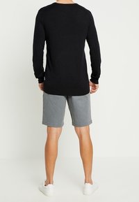 Mann in schwarzem Langarmshirt, grauen knielangen Shorts und weißen Turnschuhen, der vor einer schlichten weißen Wand steht.