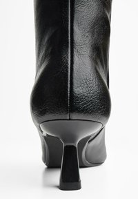 Bottine en cuir noir avec un talon noir brillant. Présente une texture lisse, un bout arrondi et des détails de couture le long de l'arrière.