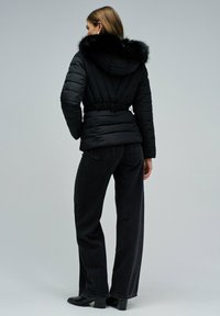 Chaqueta negra acolchada con capucha forrada de piel, cintura entallada y costuras verticales. Combinada con vaqueros negros de talle alto y botines.