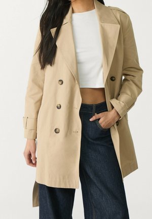 Vrouw draagt een beige double-breasted trenchcoat, wit cropped top en donkerblauwe high-waisted jeans, een hand in de zak.