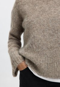 Svetlohnedý, nadýchaný sveter vyrobený z mäkkého materiálu s voľným strihom. Obsahuje dlhé rukávy a rebrovaný lem, položený na bielom tričku.