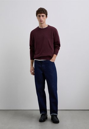 Maroon sweater with crew neck and long sleeves, paired with dark blue jeans. Model stands with one hand in pocket on a plain background.