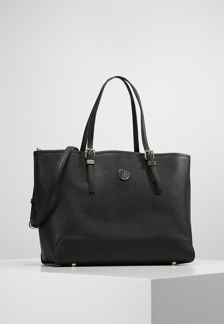 Sac cabas en cuir noir avec double poignée, bandoulière réglable, détails boucle argentée et un emblème rond en métal avec logo à l'avant.