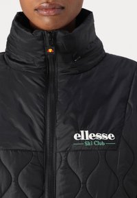 Czarna pikowana kurtka z wysokim kołnierzem, zapięciem na zamek i haftowanym logo "ellesse Ski Club" w białym i zielonym kolorze. Czerwony akcent z logo przy zamku.