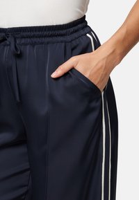 Granatowe spodnie sportowe z paskiem ściągającym w pasie, boczną kieszenią i kontrastującymi białymi paskami po bokach. Gładki, lekki materiał.