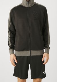 Giacca nera con zip e alto colletto, dettagli grigi e logo sul petto; abbinata a pantaloni corti neri coordinati con logo all'orlo.