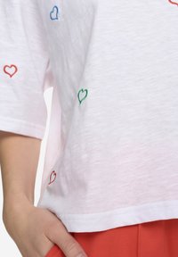 Bílé tričko s krátkým rukávem se scattered vyšívanými srdíčky v červené, zelené a modré barvě. Lehká látka s volným střihem.
