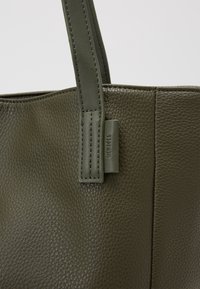 Sac en cuir vert olive avec une finition texturée, de forme incurvée et des poignées assorties ; il possède une petite étiquette de marque sur le côté.