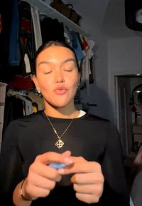 Jeune personne aux yeux fermés tenant un petit objet, debout devant un portant à vêtements dans une pièce faiblement éclairée.