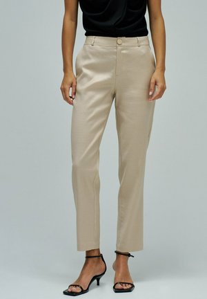 Femme portant un pantalon beige sur mesure et des sandales à talons noires à brides, debout devant un fond uni gris clair.