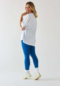 Biała, oversize'owa koszulka, niebieskie legginsy, białe skarpetki i beżowe buty sportowe. Gładki materiał, luźny krój i subtelne detale designu.