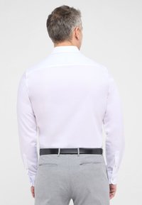 Witte blouse met lange mouwen en een gladde textuur, rugpasdetail en contrasterende knoopaccenten. Gecombineerd met lichtgrijze broek en een zwarte riem.