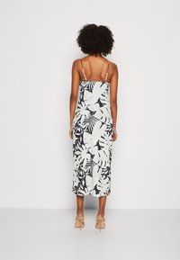 Femme de dos portant une robe midi à imprimé tropical noir et blanc avec des fines bretelles spaghetti et des talons nus.