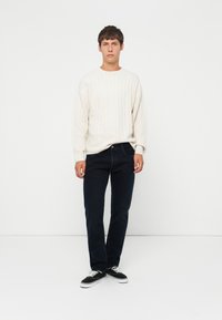 Crème ribgebreide trui, donkerblauwe jeans, zwarte sneakers, staand tegen een witte achtergrond, met een eenvoudige, minimalistische uitstraling.