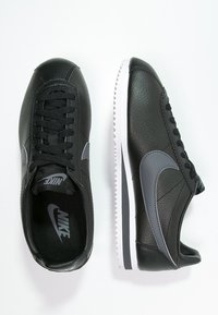 Chaussures de sport en cuir noir avec des accents gris et des lacets. Le design présente un logo swoosh distinctif, une tige texturée, et une semelle blanche.