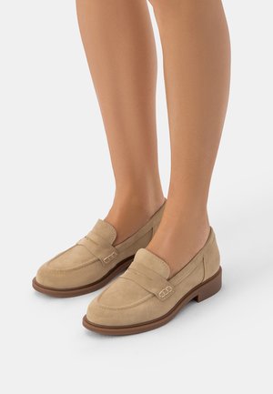 Nogi w beżowych zamszowych mokasynach na niskim obcasie z przeszyciami na białym tle.