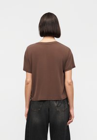 Άτομο με κοντά σκούρα μαλλιά που φοράει απλό καφέ κοντομάνικο πουκάμισο και ξεθωριασμένο μαύρο τζιν με φαρδιά μπατζάκια, με την πλάτη στραμμένη προς τα πίσω πάνω σε λευκό φόντο.